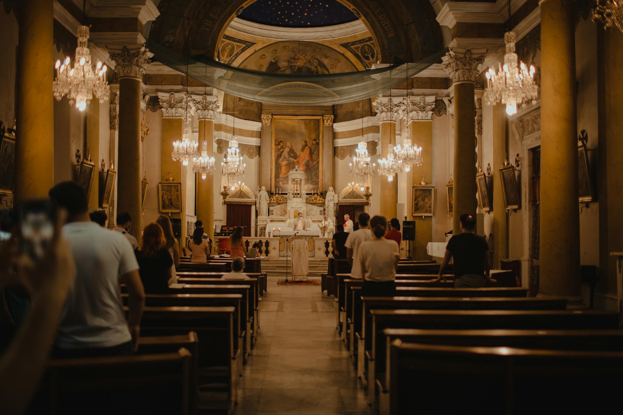 Interior view of a Catholic church with chandeliers and people attending a service.